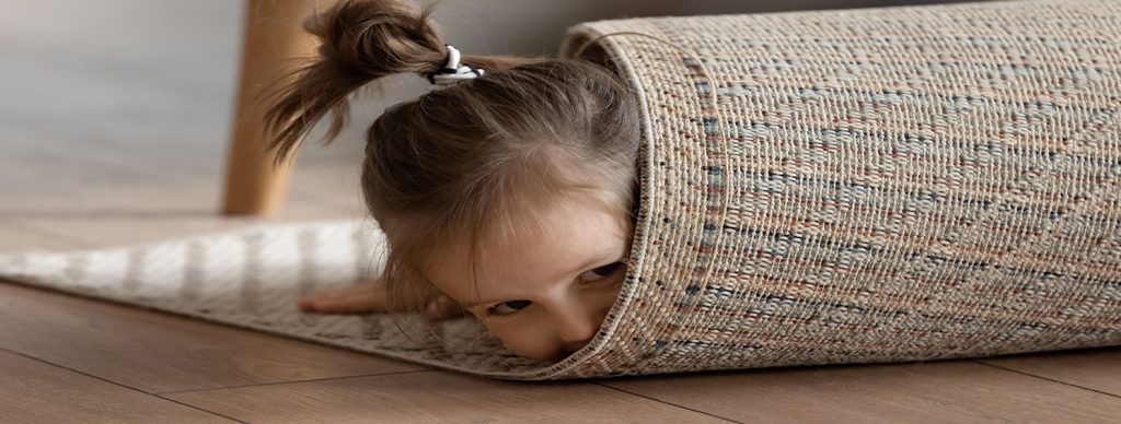 Adorable small girl wrapped in rug lying on warm wooden laminate floor in modern living room alone, close up view. Playtime funny hiding games, professional cleaning carpet company services ad concept